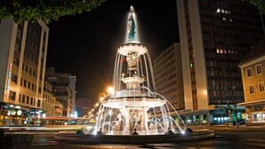 Une vue nocturne d’une fontaine à plusieurs niveaux, avec des jets d’eau, entourée de bâtiments modernes et d’enseignes lumineuses.