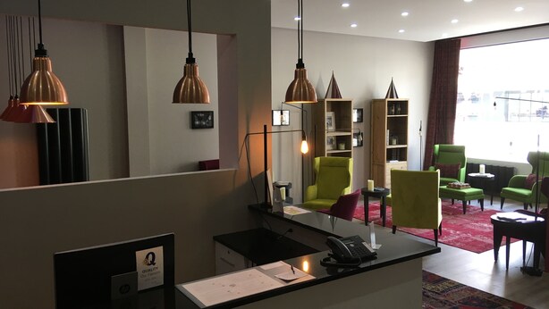 Un espace d’accueil d’hôtel avec un comptoir noir, un téléphone, un panneau portant la lettre « Q » et une grande fenêtre donnant sur un paysage.