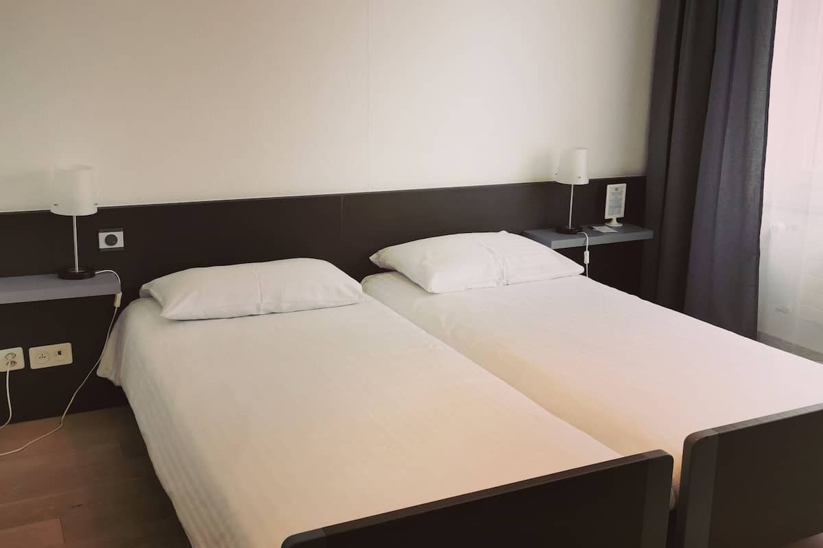 Uma cama de casal com roupa de cama branca, cabeceira escura, mesas de cabeceira com abajures e piso de madeira.