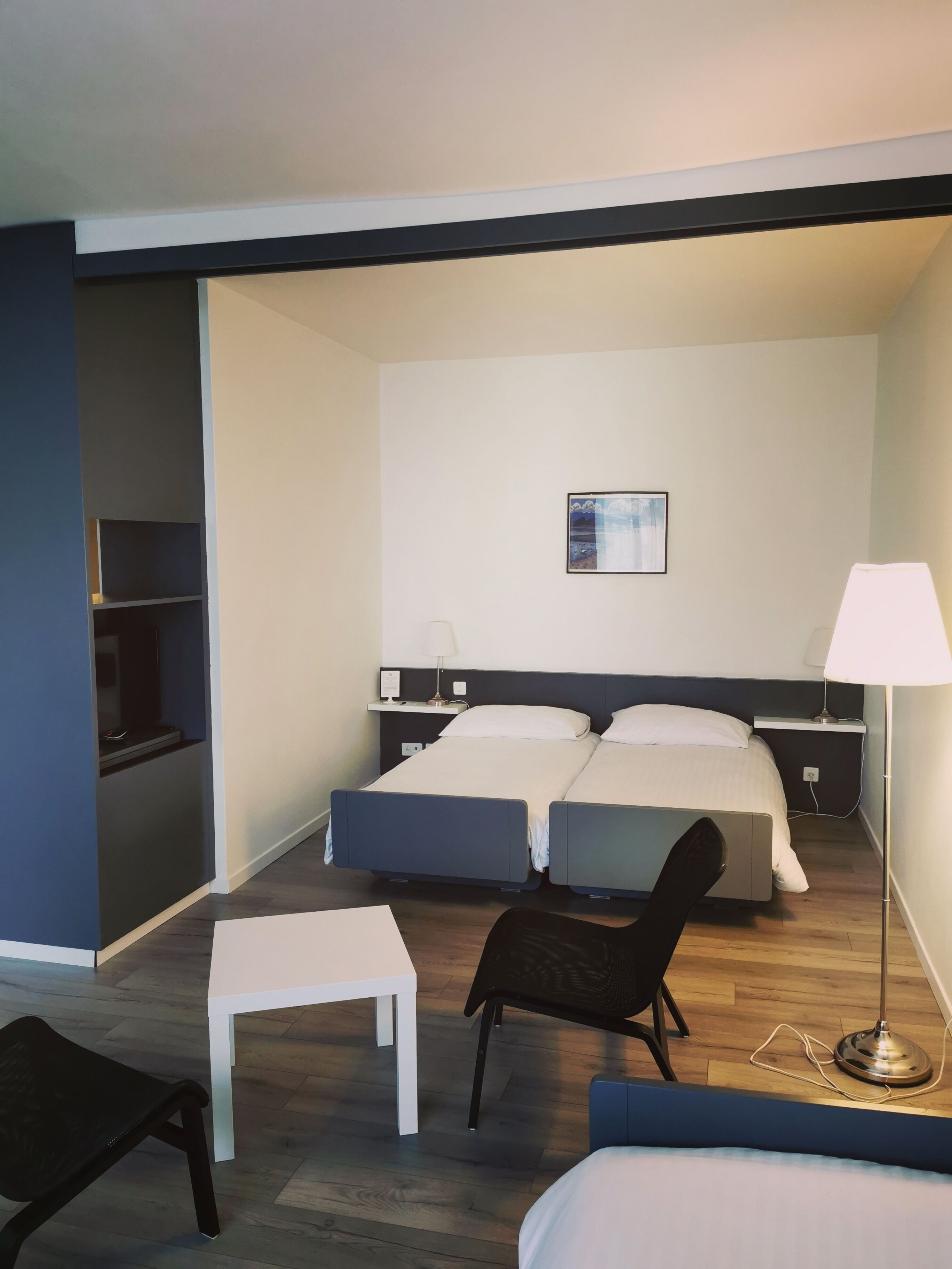 Habitación de hotel con dos camas, un escritorio, una silla, una lámpara de pie y un estante pequeño.