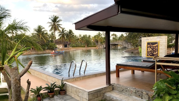 A swimming pool with a lounge area, surrounded by palm trees and a stone bridge. - Rayong Rental Pool Villas (Klaeng)