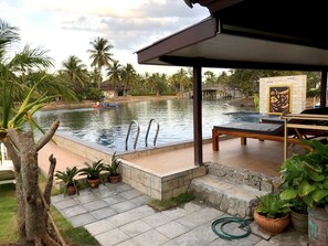Una piscina con zona de descanso, rodeada de palmeras y un puente de piedra.