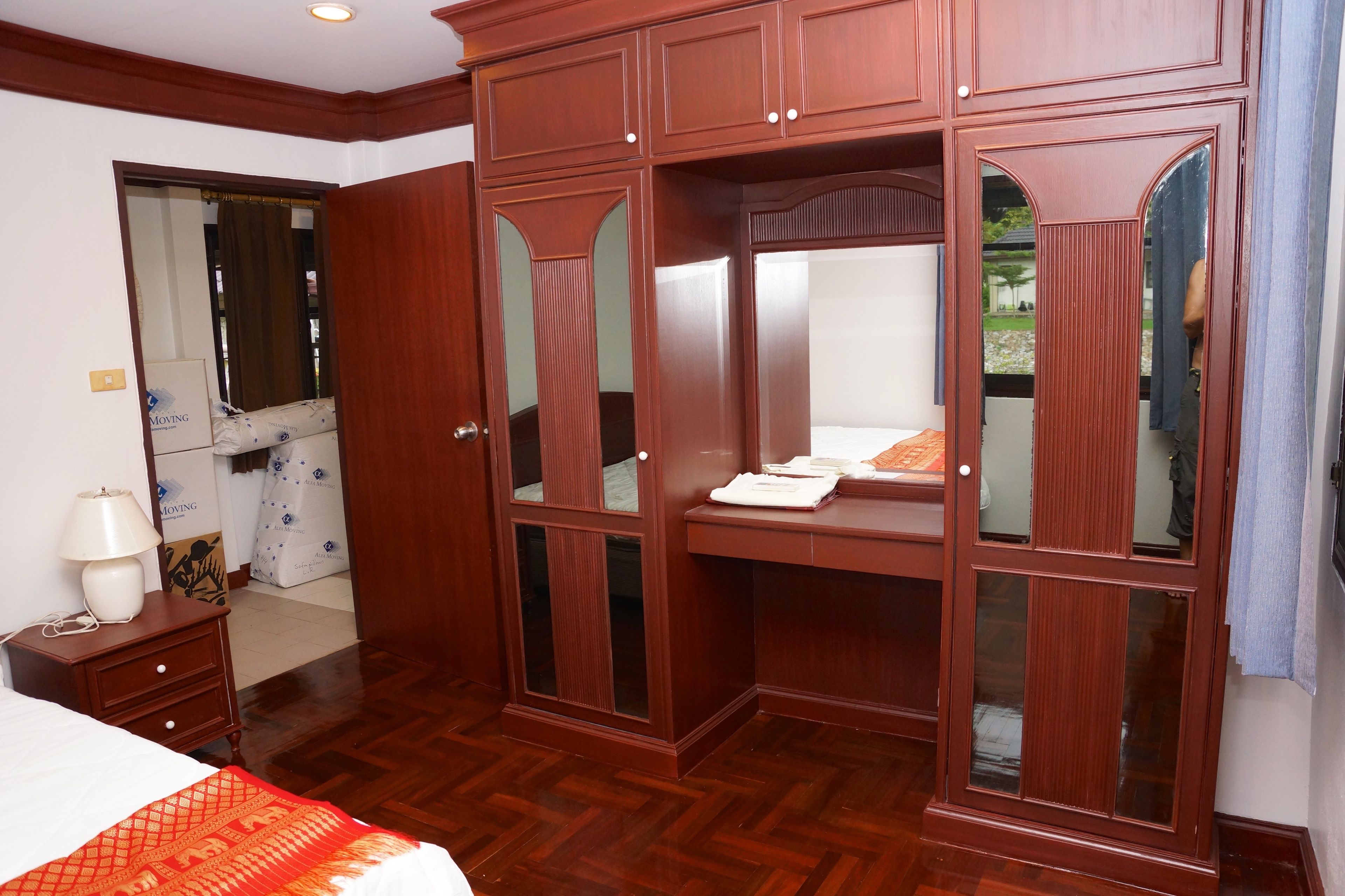 A bedroom with a wooden wardrobe, a bed with a patterned bedspread, a nightstand with a lamp, and a mirror.