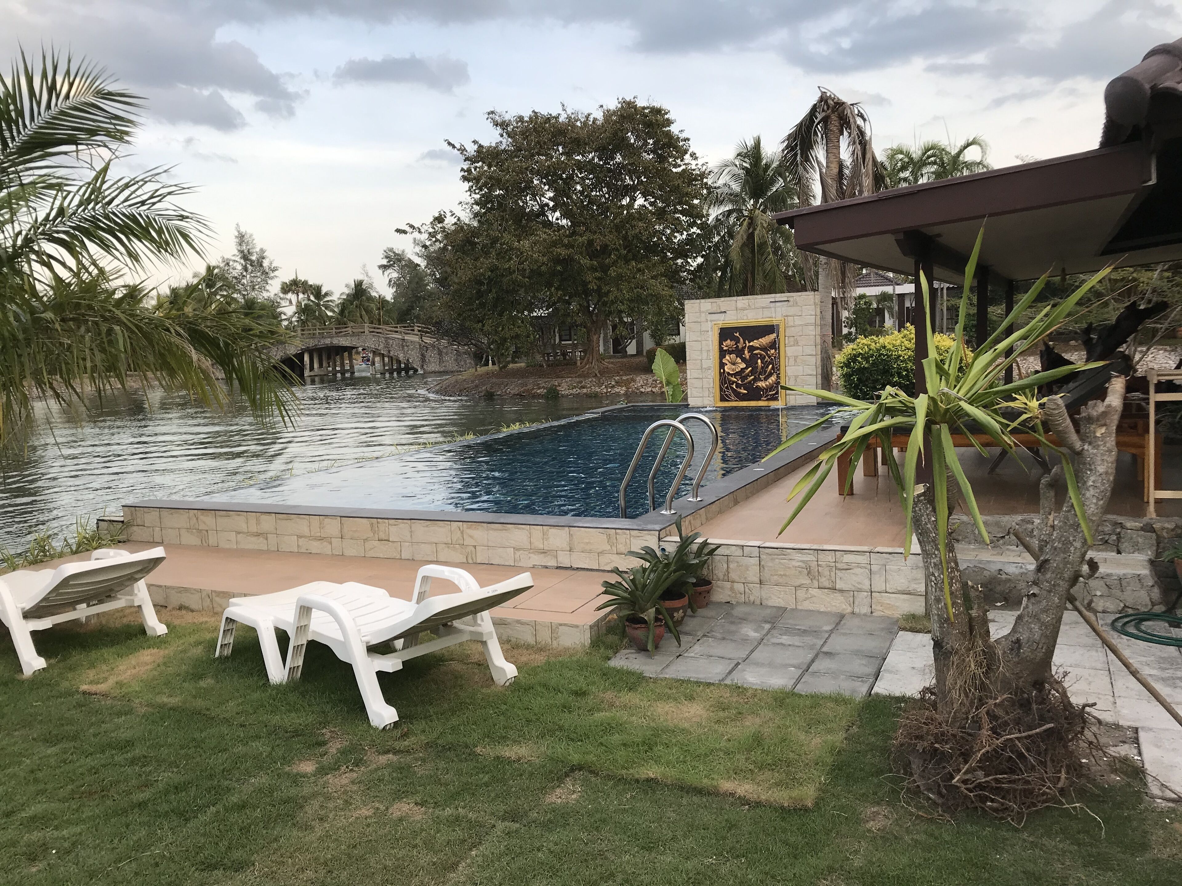 A swimming pool with a stone border, two white lounge chairs, and a small bridge over a water feature.