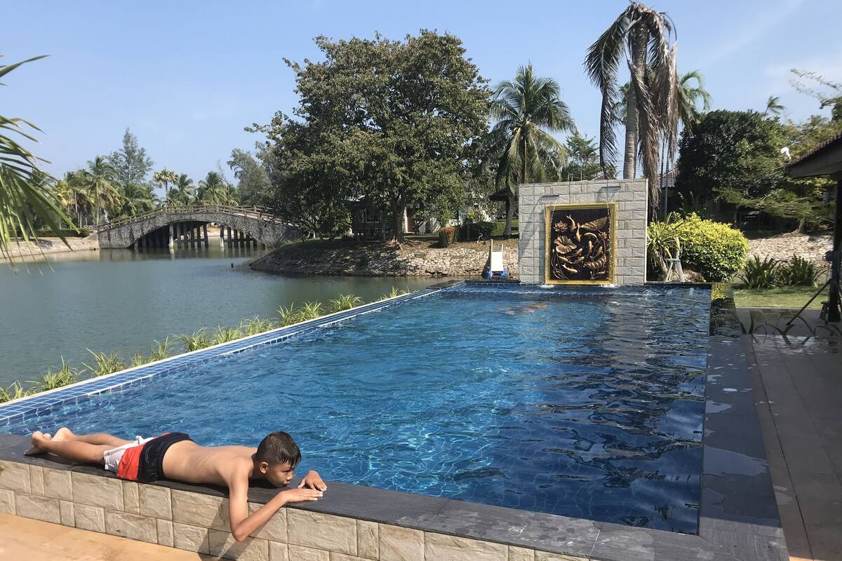 A swimming pool with a stone border, a bridge, and palm trees in the background.