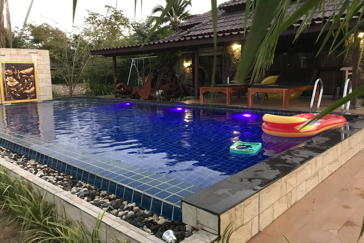 A swimming pool with a tiled edge, surrounded by a stone wall and a small garden area with rocks and plants.