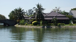 A traditional house with a tiled roof, surrounded by palm trees and a stone bridge over a calm body of water. - Rayong Rental Pool Villas (Klaeng)