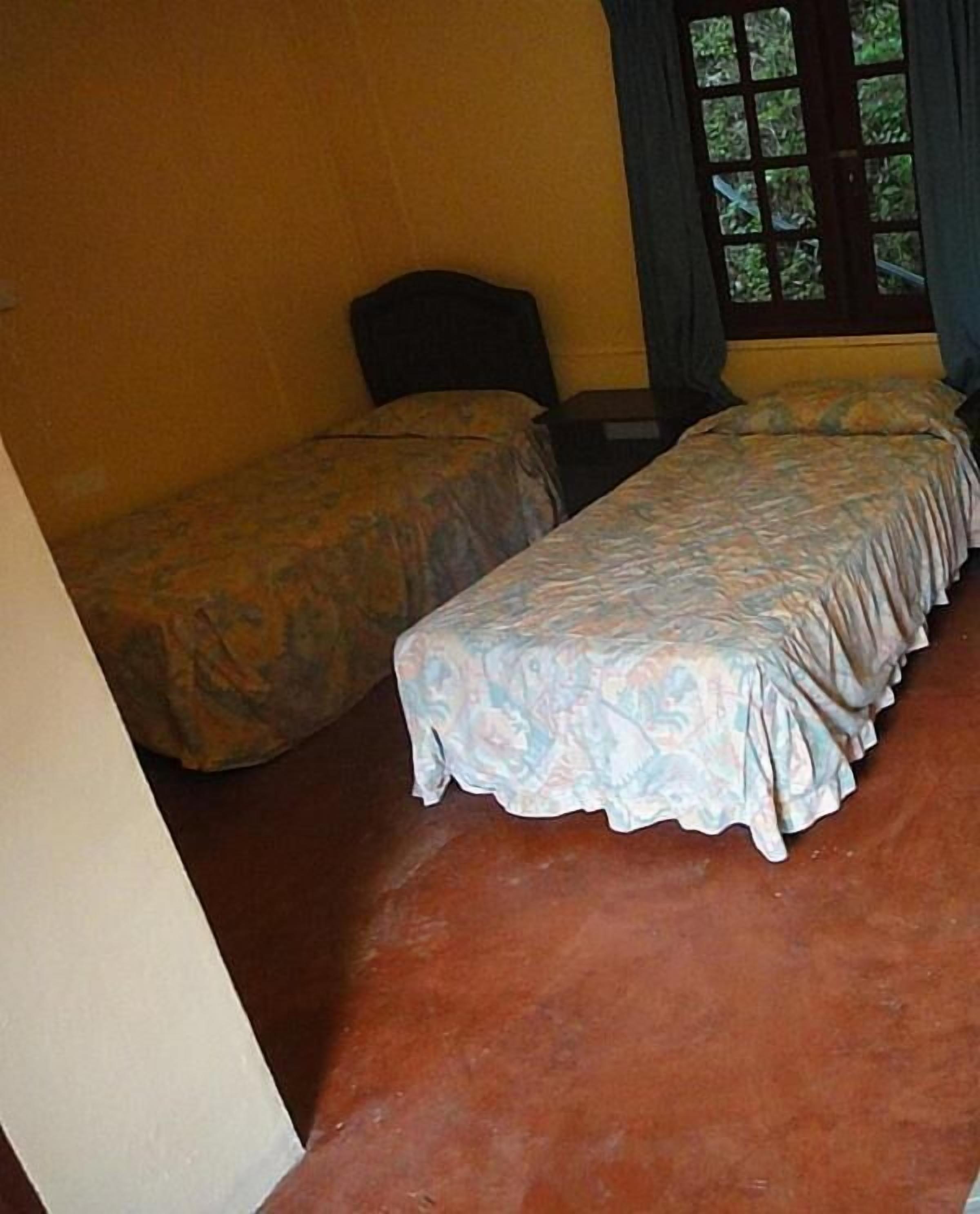 A room with two beds, a window with a view of greenery, and a brown floor.