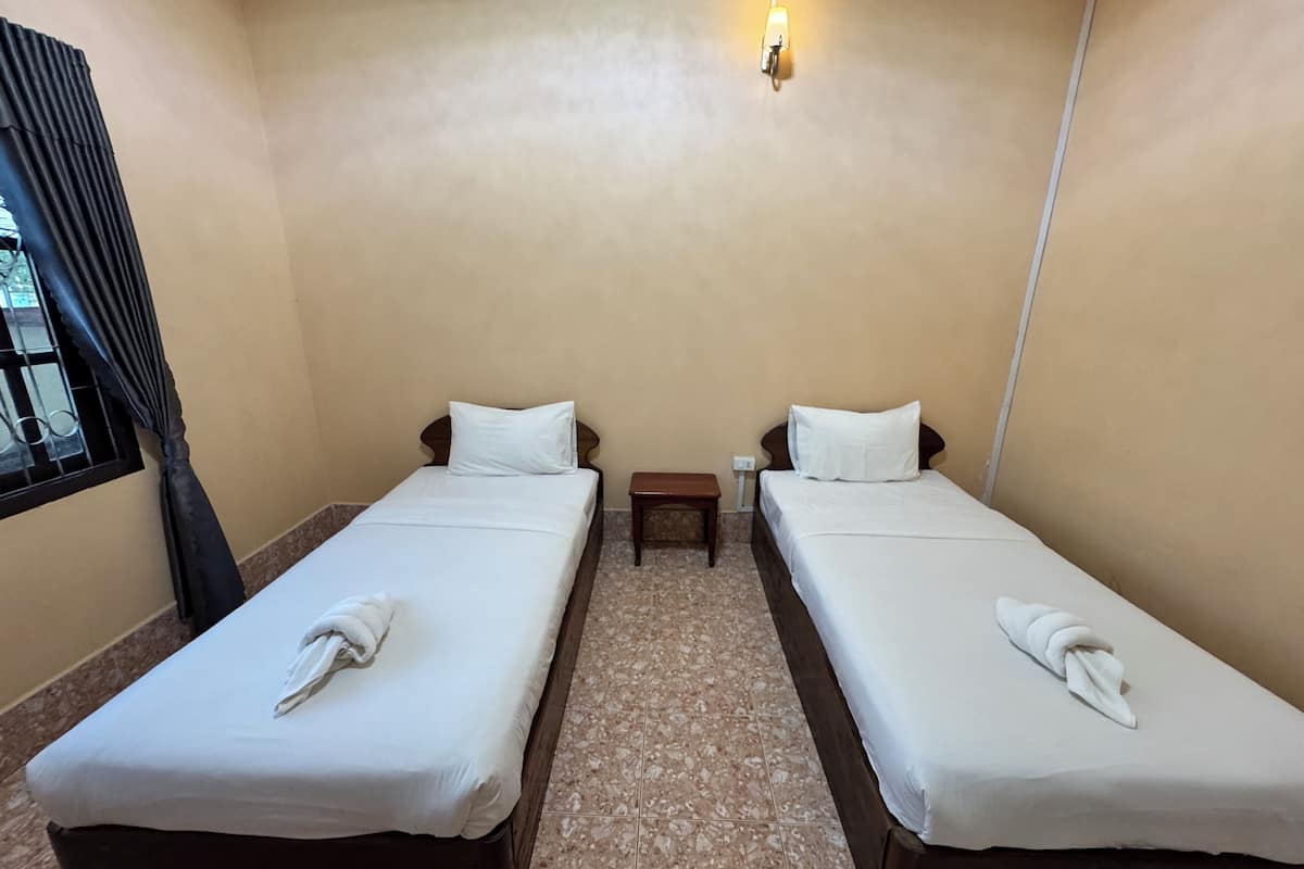 Quarto de hotel com duas camas de solteiro, cada uma com um lençol branco e uma toalha. Há uma pequena mesa de cabeceira de madeira entre as camas, e um luminário fixado na parede acima delas.