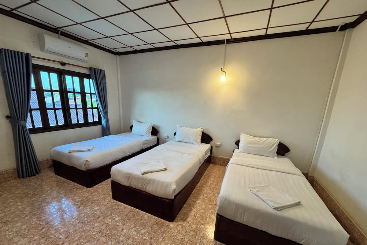 Quarto de hotel com três camas simples, uma janela com cortinas, ar condicionado e piso ladrilhado.
