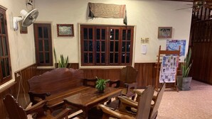 Un coin repas traditionnel, avec une grande table et des chaises en bois, un ventilateur fixé au mur et un tableau d’affichage où sont accrochés divers documents.