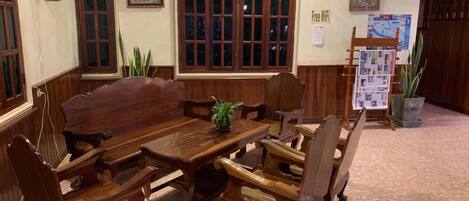 Un coin repas traditionnel, avec une grande table et des chaises en bois, un ventilateur fixé au mur et un tableau d’affichage où sont accrochés divers documents.