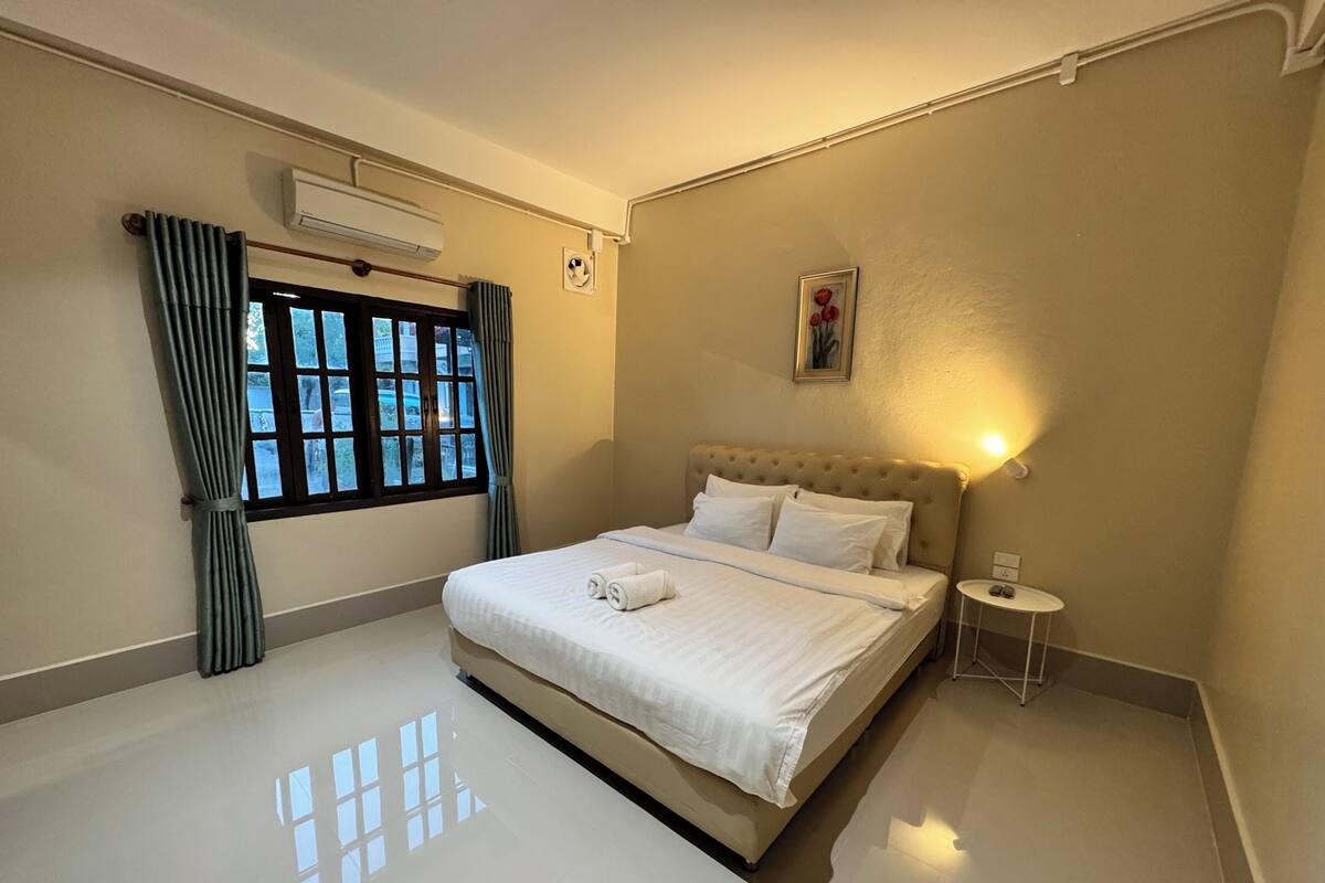 Quarto de hotel com cama grande, mesa de cabeceira pequena, janela com cortinas, aparelho de ar condicionado e luminária fixada na parede.