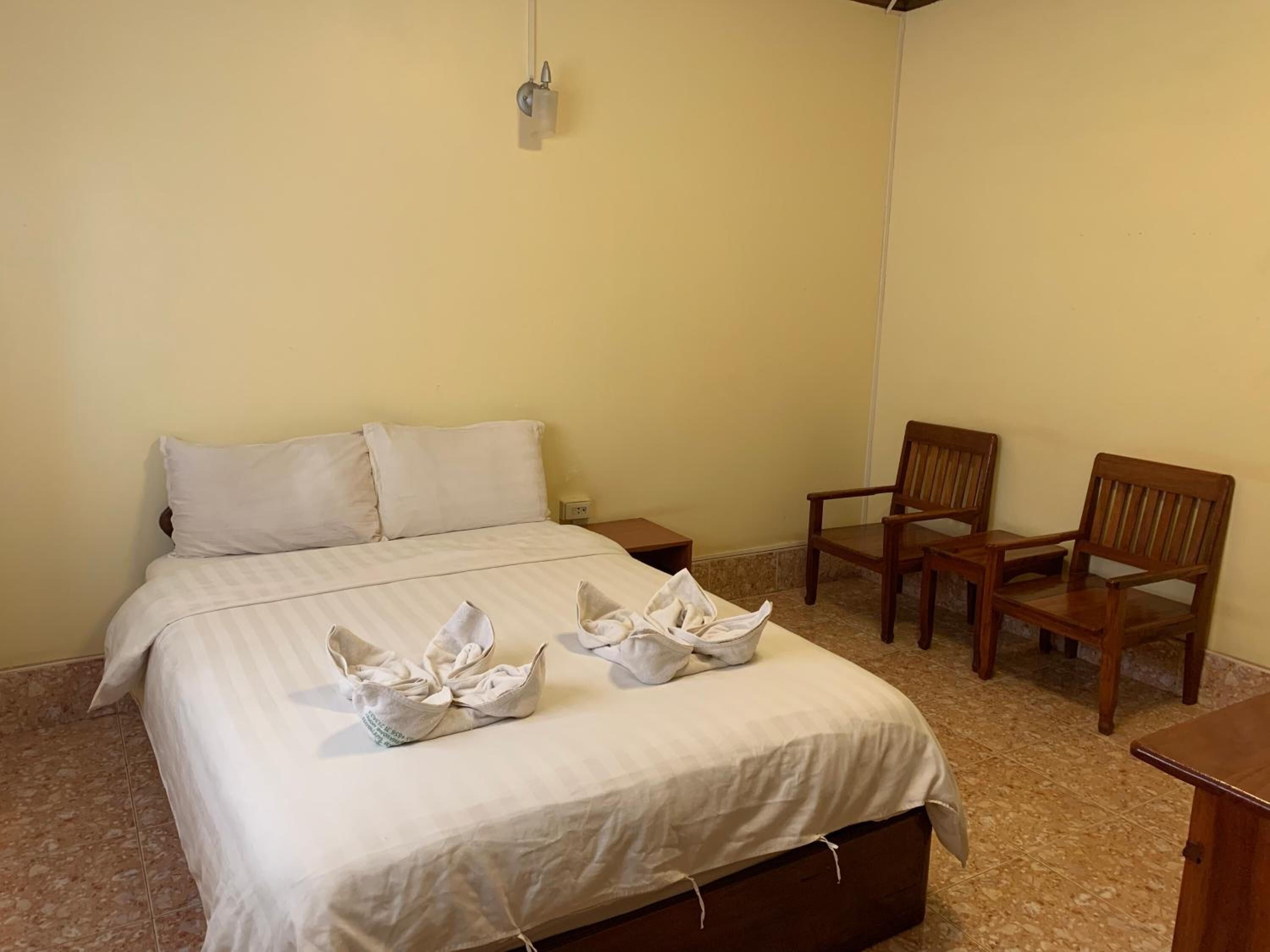 A neatly made bed with a white bedspread and pillows, two wooden chairs, and a small bedside table.