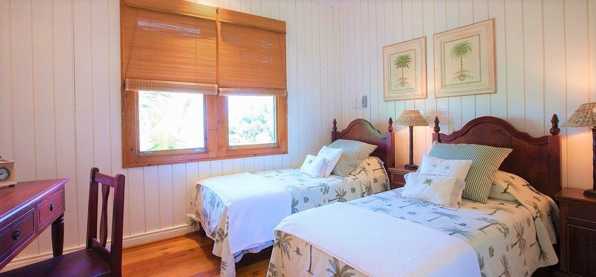 A bedroom with two single beds, a wooden desk, and a chair. There are two bedside tables with lamps, a window with blinds, and framed pictures on the wall.