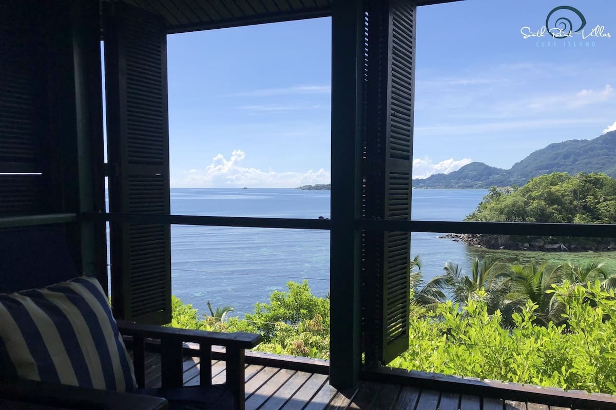 Ein Zimmer mit einer Holzterrasse und Blick auf ein Gewässer, eine üppige grüne Landschaft und einen klaren Himmel.
