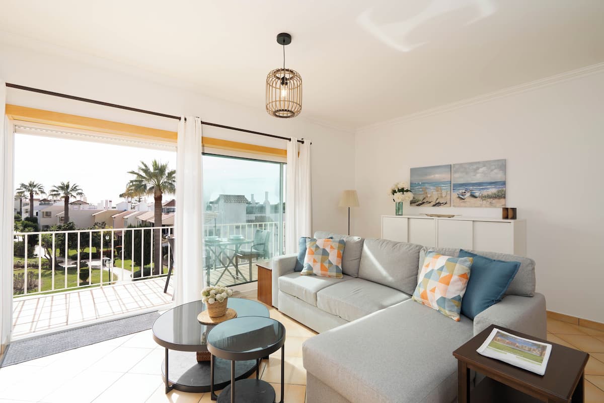A modern living room with a grey sofa, two side tables, and a balcony with a view of palm trees and buildings.