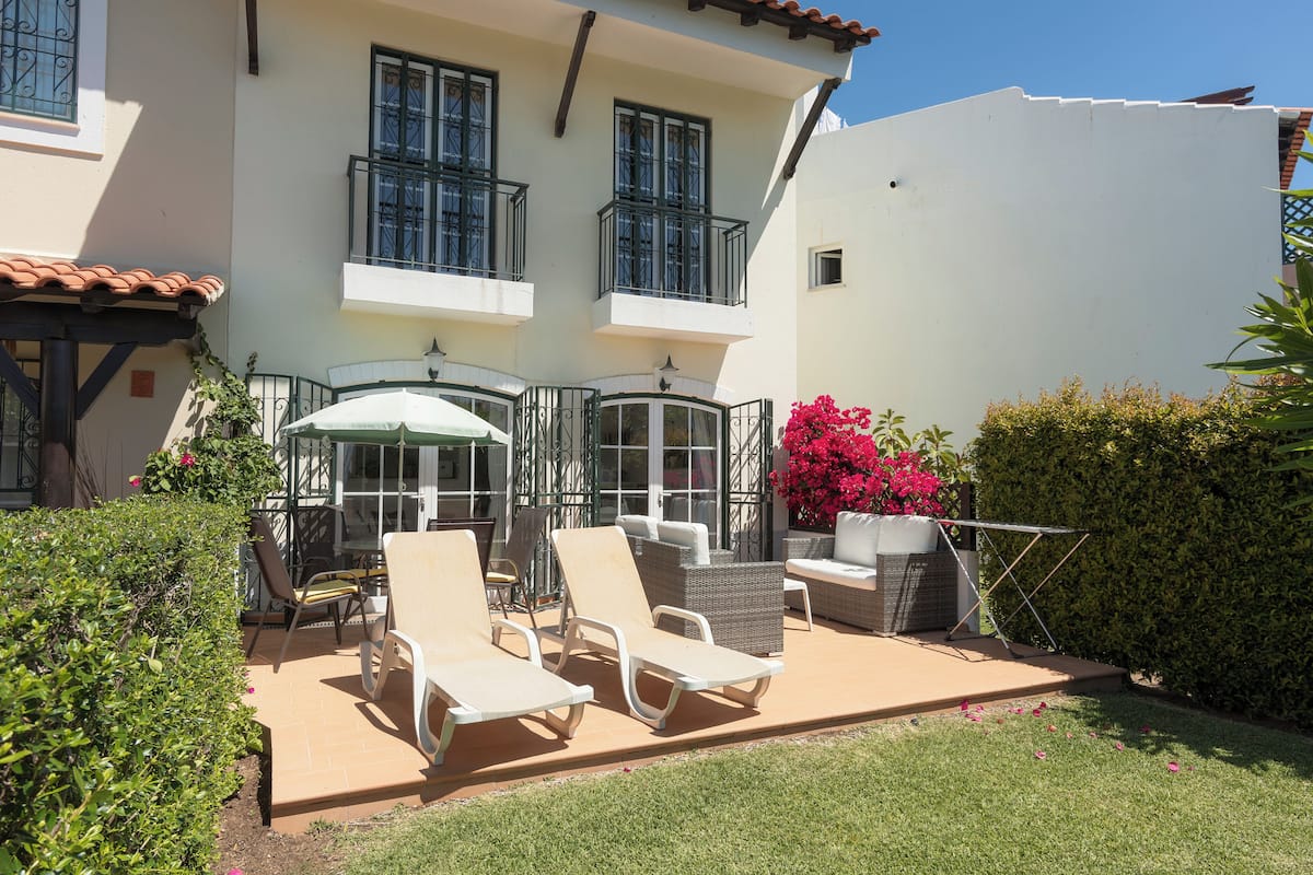 A patio with sun loungers and a table, surrounded by a white building with balconies and a green hedge.