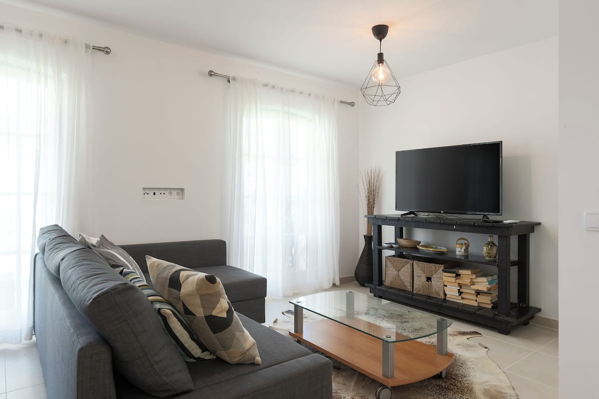 A modern living room with a grey sofa, a glass coffee table, a black entertainment unit with books, and a flat-screen TV.