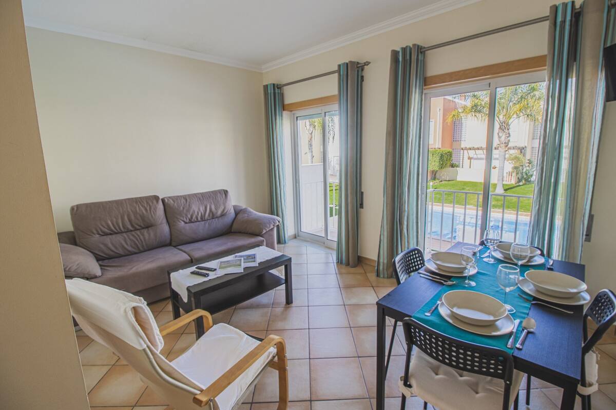A living area with a sofa, a dining table set for two, and a view of a pool and garden through large windows.