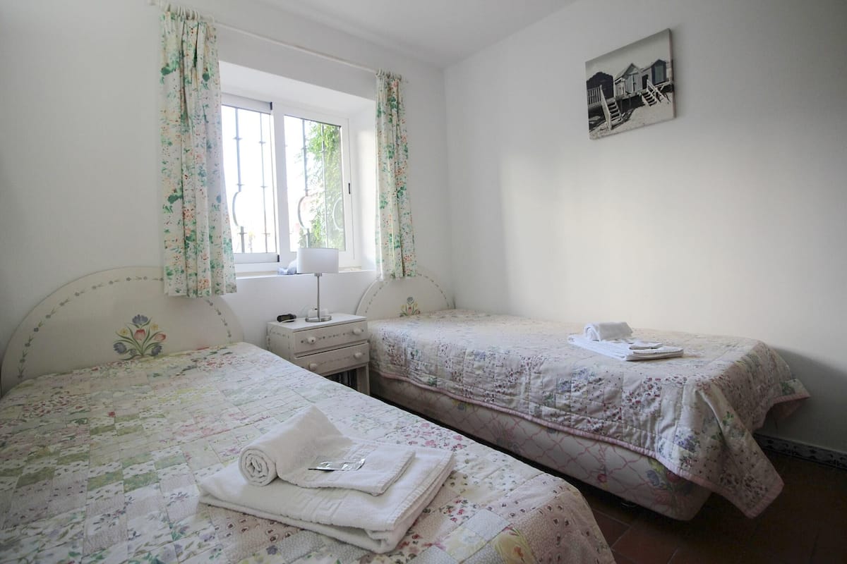 A bedroom with two beds, a window with curtains, a bedside table with a lamp, and a framed picture on the wall.