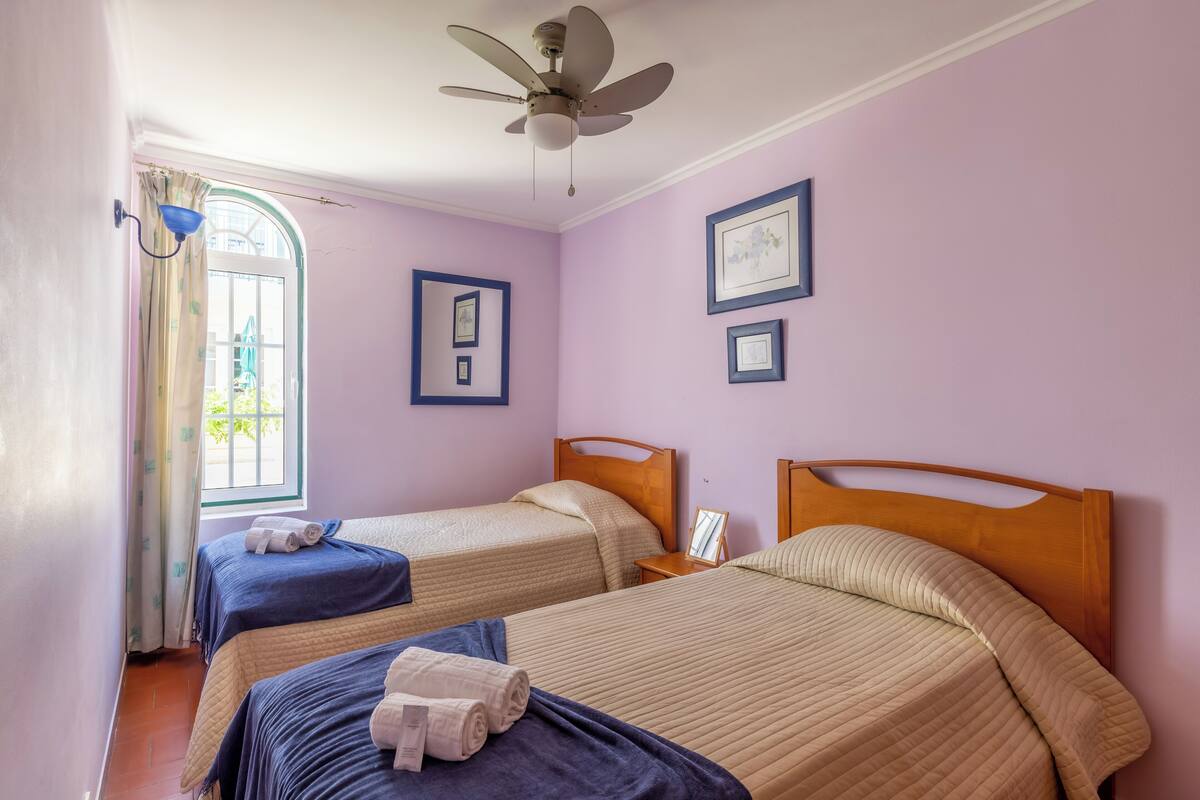 A room with two beds, a ceiling fan, and framed pictures on the walls.