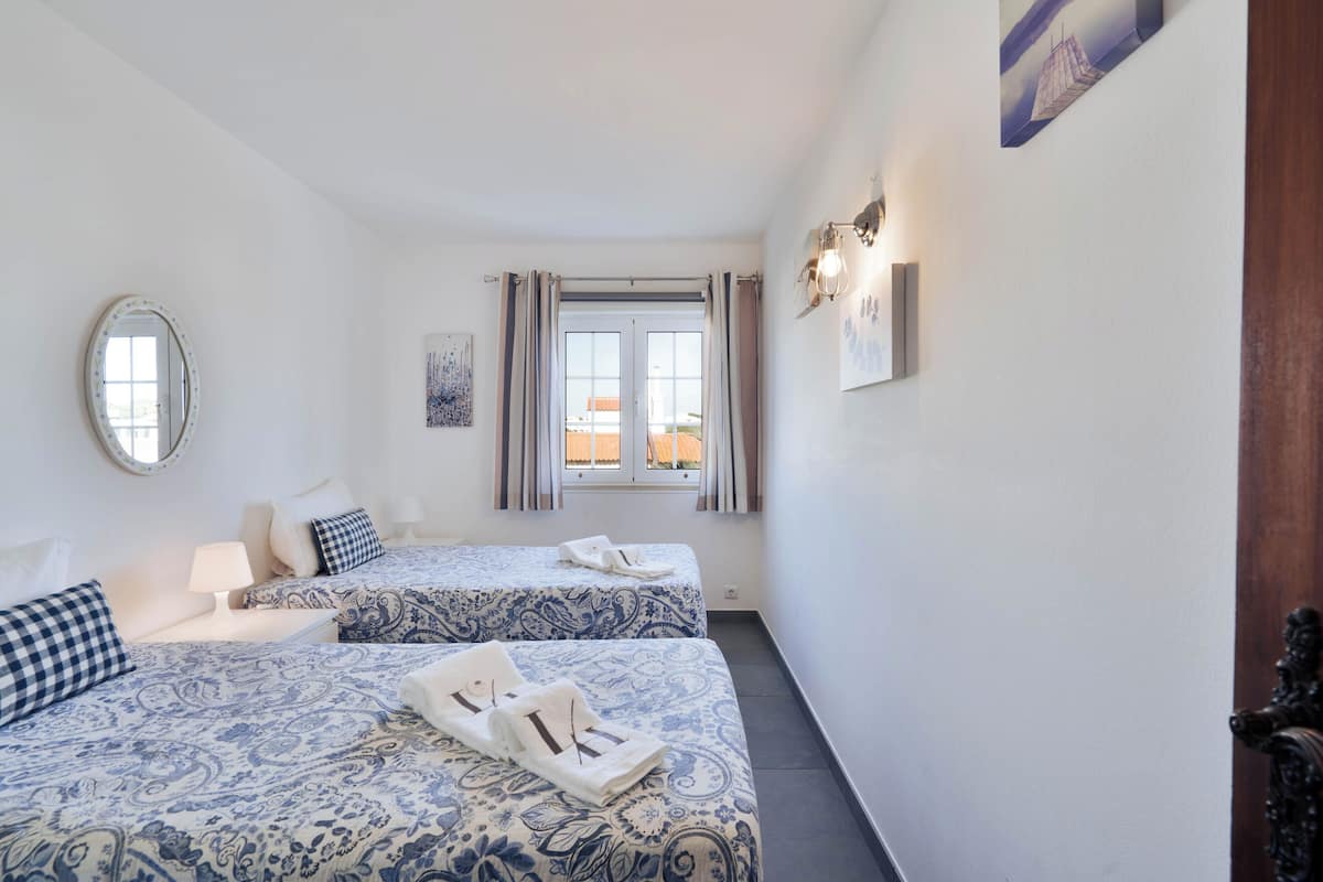 A bedroom with two beds, a window with curtains, a wall-mounted light fixture, and a mirror.