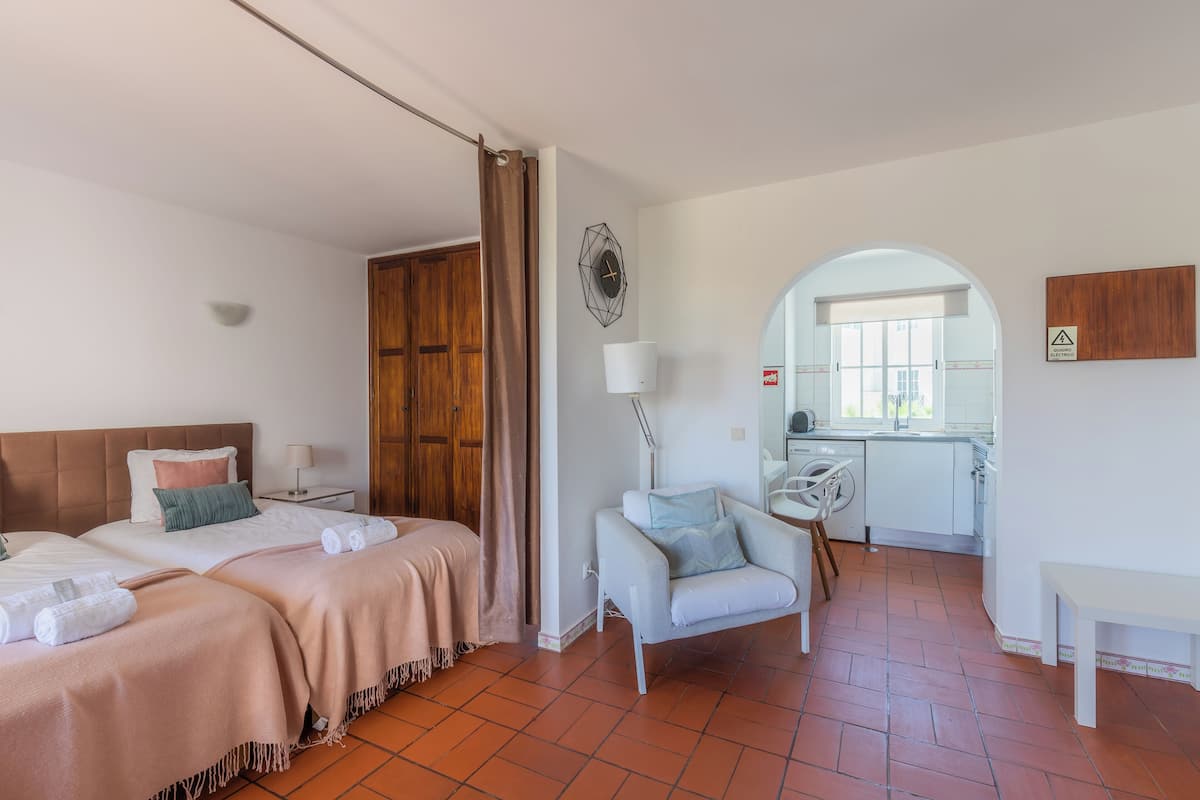A hotel room with two beds, a sitting area with a chair, a small table, and a view of the kitchen and balcony.