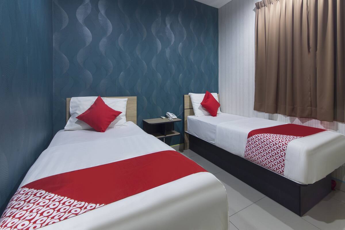 Quarto de hotel com duas camas, cada uma com roupa de cama branca e detalhes vermelhos. Tem uma mesa de cabeceira com telefone e luminária. O quarto tem uma parede com padrão azul e uma janela com cortinas marrons.