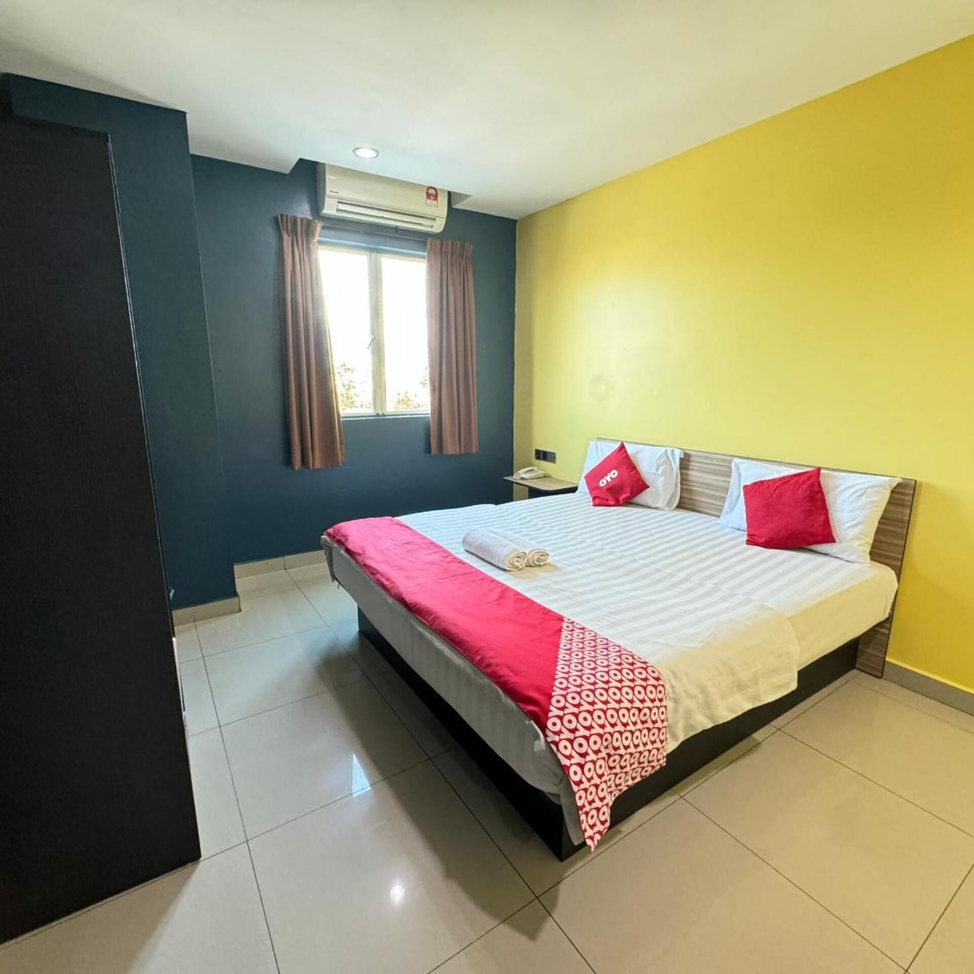 A hotel room with a bed, a nightstand, a window with curtains, and an air conditioning unit.