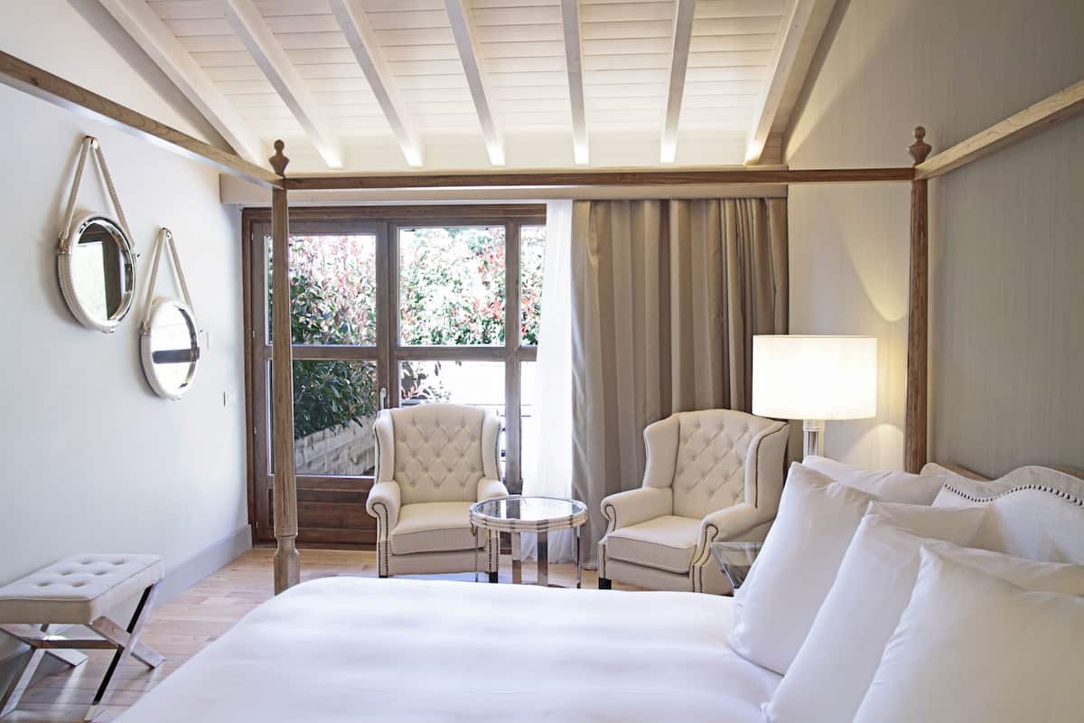 Een hotelkamer met een groot bed, twee fauteuils, een klein rond tafeltje en een raam met gordijnen.