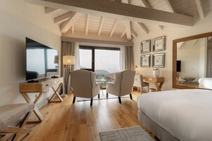 Habitación de hotel con una cama grande, un escritorio con televisor de pantalla plana, dos sillones, un ventanal con vistas y un techo con vigas a la vista.