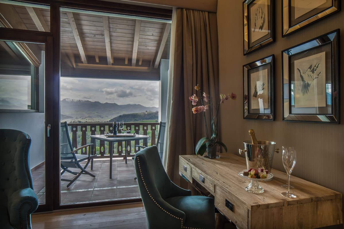 Een kamer met een houten tafel, een met groen bekleden stoel, een balkon met uitzicht op bergen en kunstwerken in lijsten aan de muur.