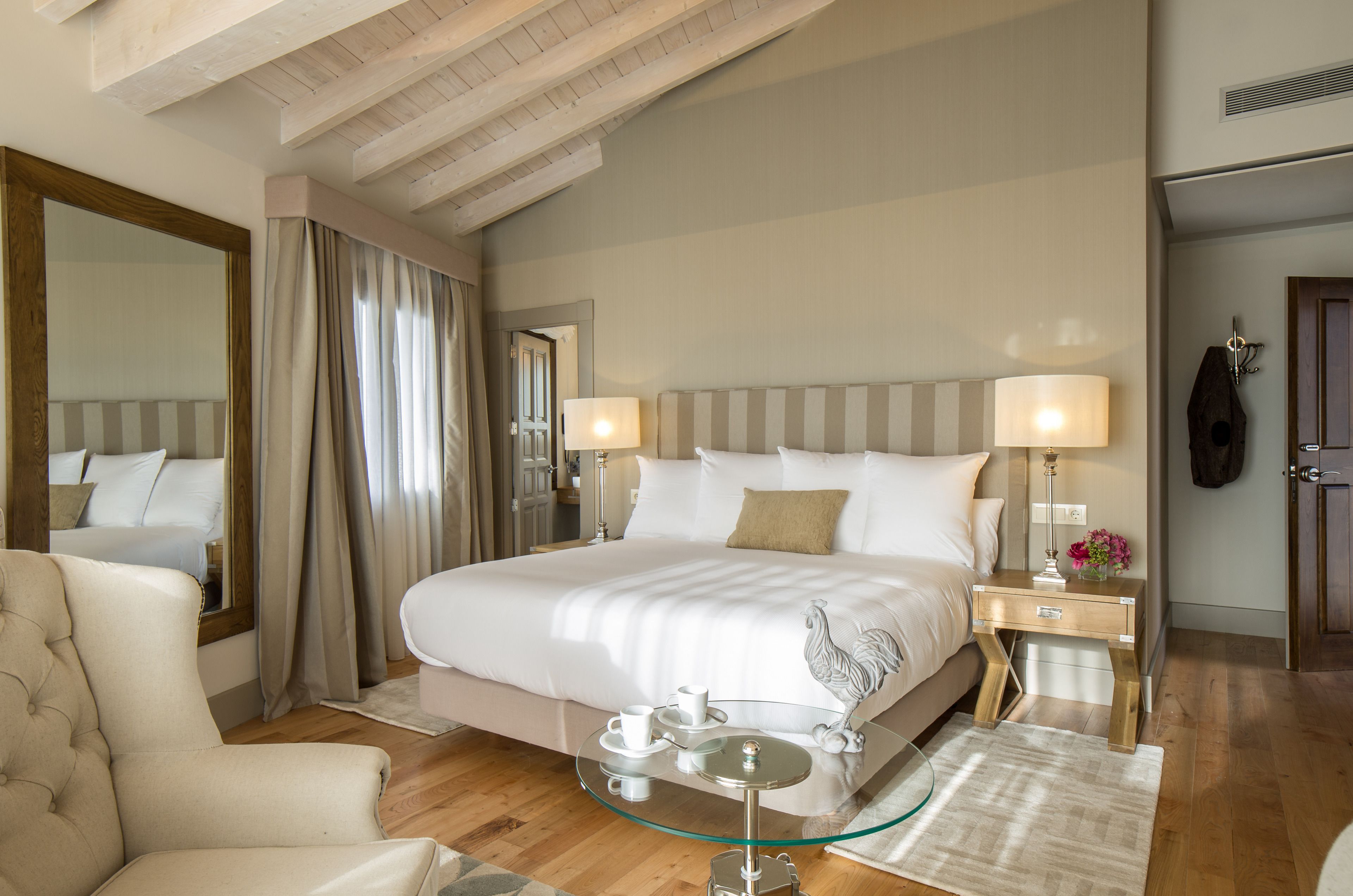 A hotel room with a large bed, two bedside tables with lamps, a glass coffee table, a chair, and a mirror.