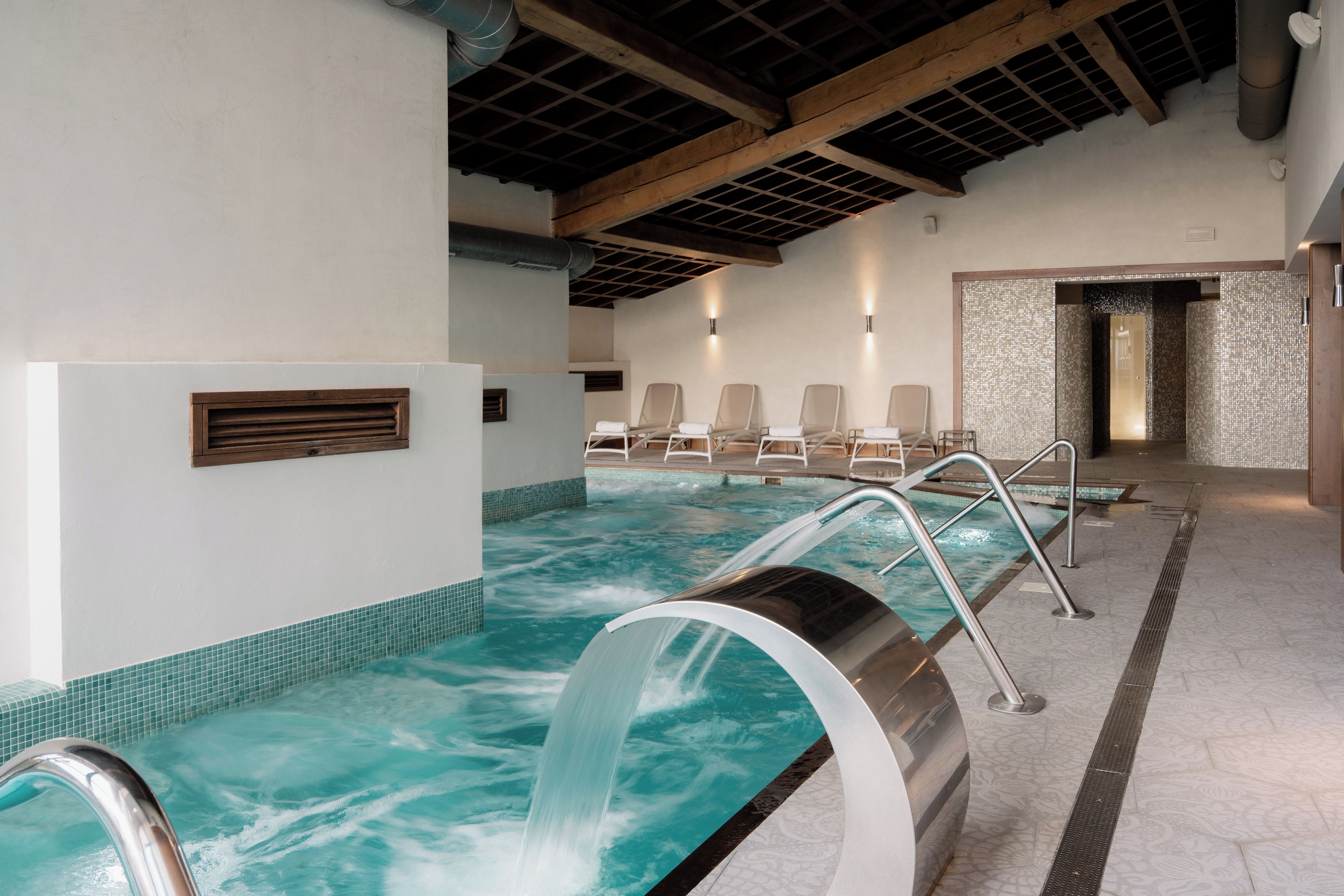 A pool area with a clear water feature, lounge chairs, and a tiled floor.