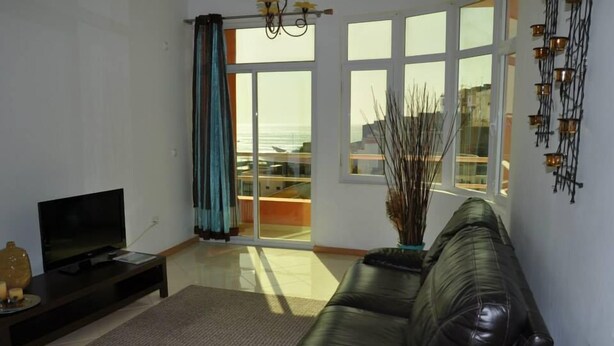 A living room with a black leather sofa, a flat-screen TV on a stand, a large window with curtains, and a view of buildings outside. - Miradouro de Gamboa (Praia)