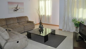 A living room with a beige sofa, a glass coffee table, a TV on a wooden stand, and a painting on the wall. - Miradouro de Gamboa (Praia)