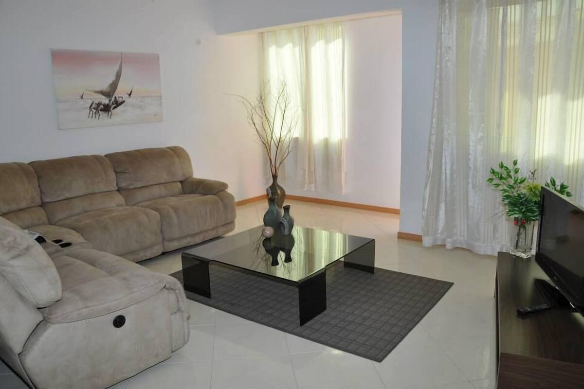 A living room with a beige sofa, a glass coffee table, a TV on a wooden stand, and a painting on the wall.