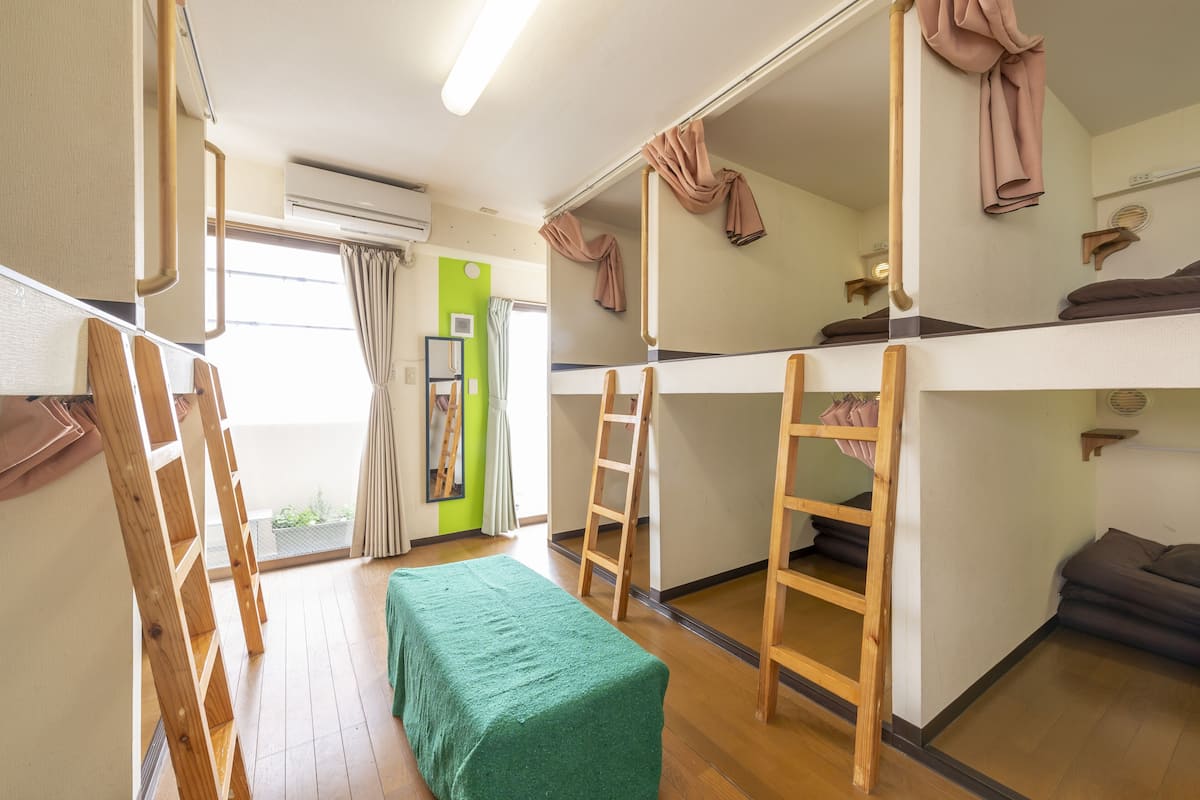 Een slaapzaal met stapelbedden, een groene tafel, houten ladders en een raam met gordijnen.