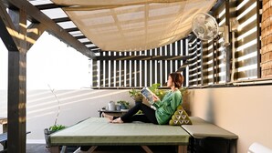 Eine Person sitzt auf einer Matte im Schatten, mit einem Ventilator, und liest ein Buch. Übertöpfe mit Pflanzen und Meerblick sind ebenfalls zu sehen.