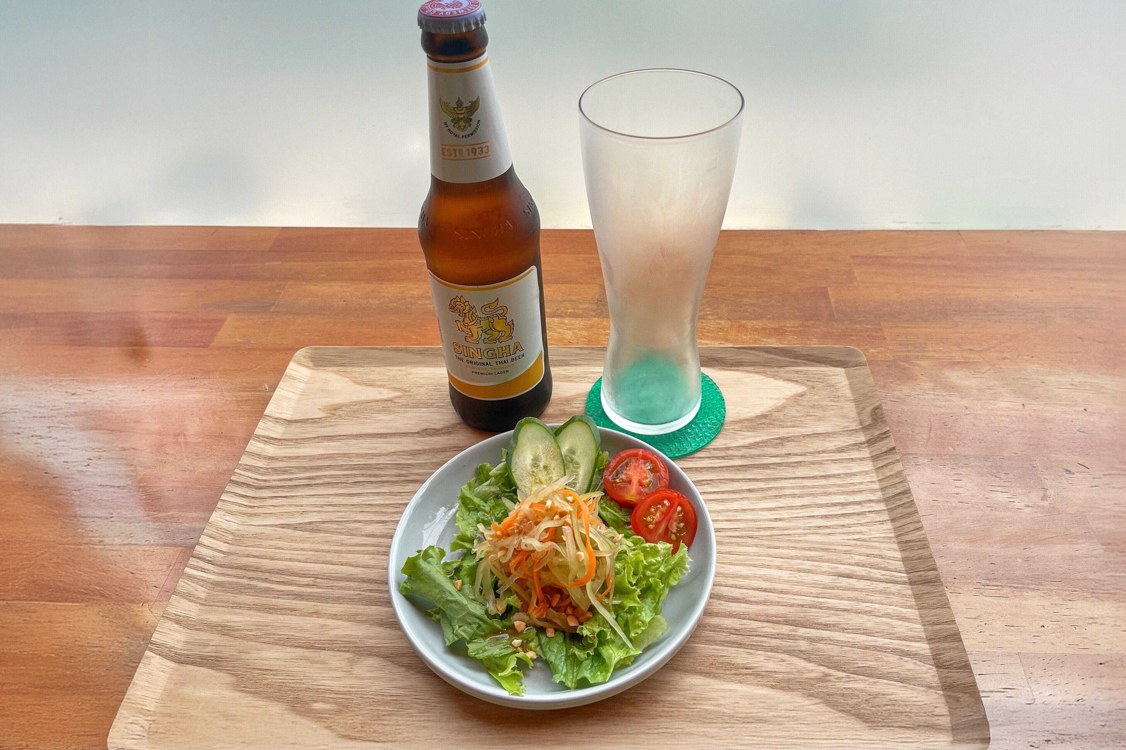 A bottle of Singha beer, a glass of water, and a plate of salad with cucumbers and tomatoes on a wooden table.