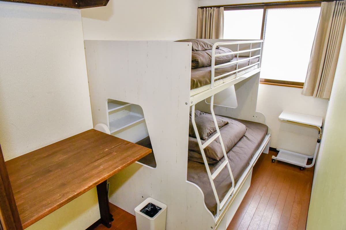 Een slaapkamer met een stapelbed, een houten plank, een kleine prullenbak en een raam met gordijnen.