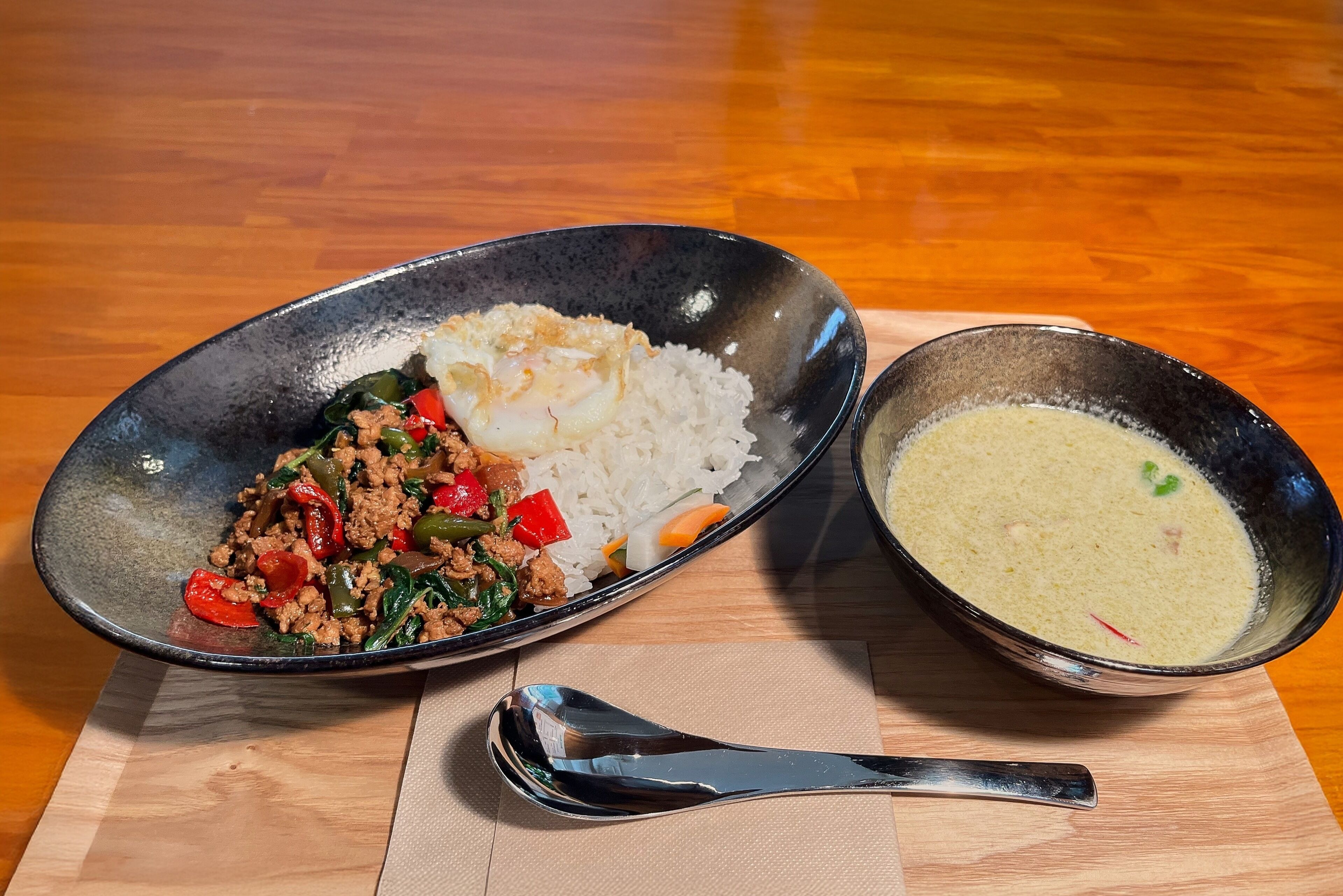 A meal consisting of stir-fried meat and vegetables, white rice, and a bowl of soup on a wooden table.