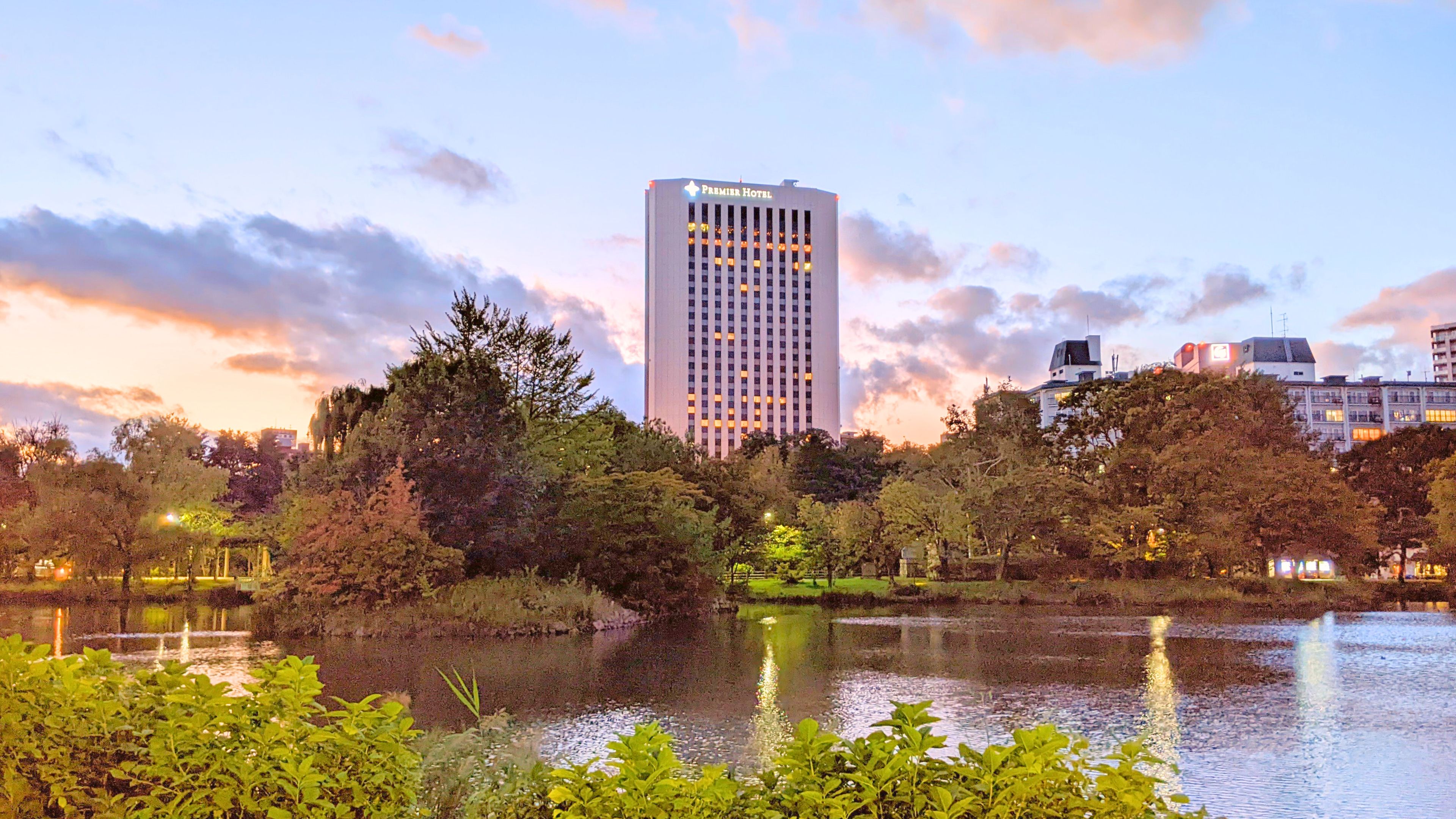A hotel with a modern design, surrounded by a park with a lake, trees, and a sunset sky.