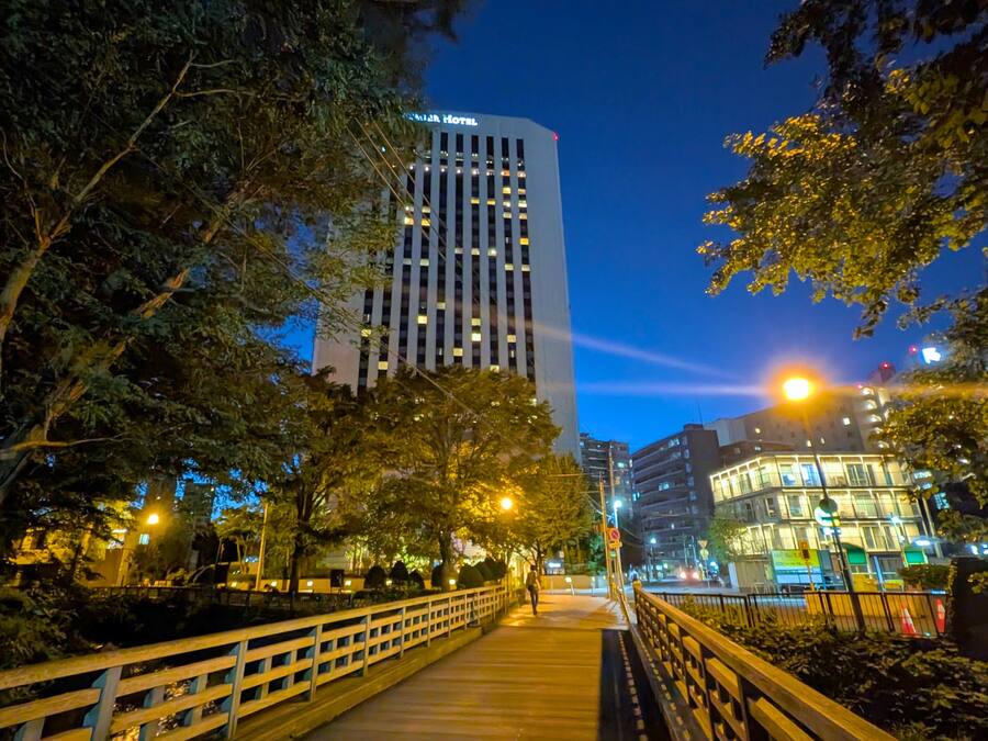 A nighttime view of a cityscape with a prominent hotel building, a pedestrian bridge, and surrounding trees.