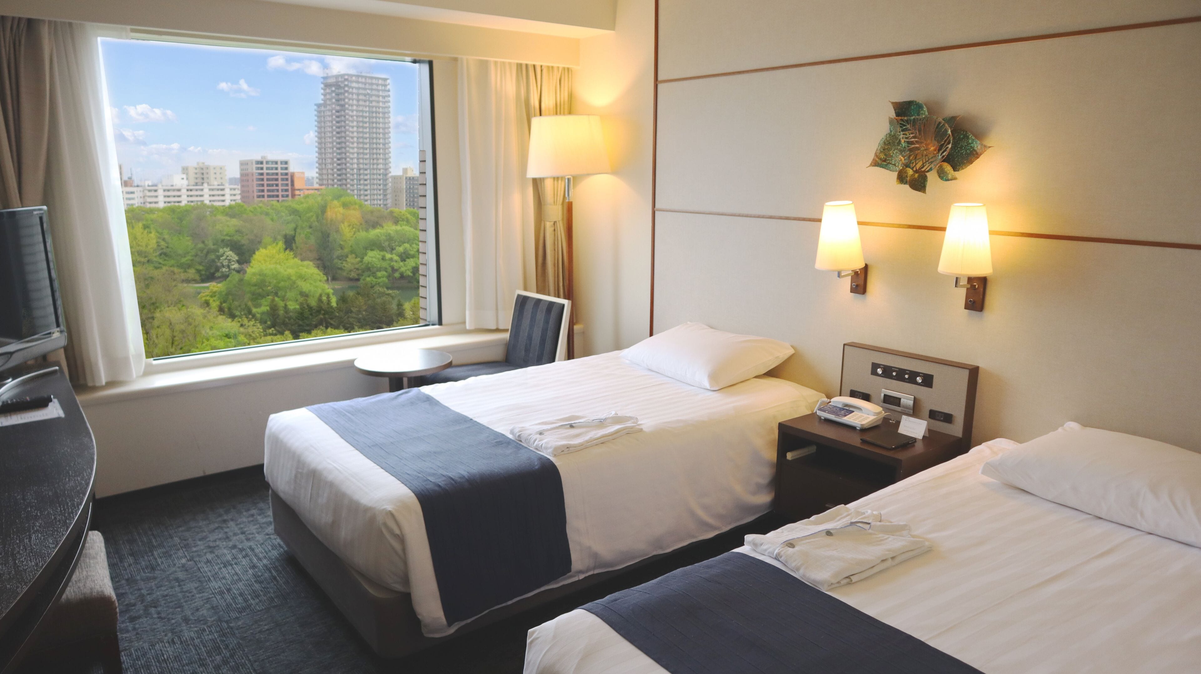 A hotel room with two beds, a desk, a chair, a small table, a window with a city view, and a wall-mounted phone.