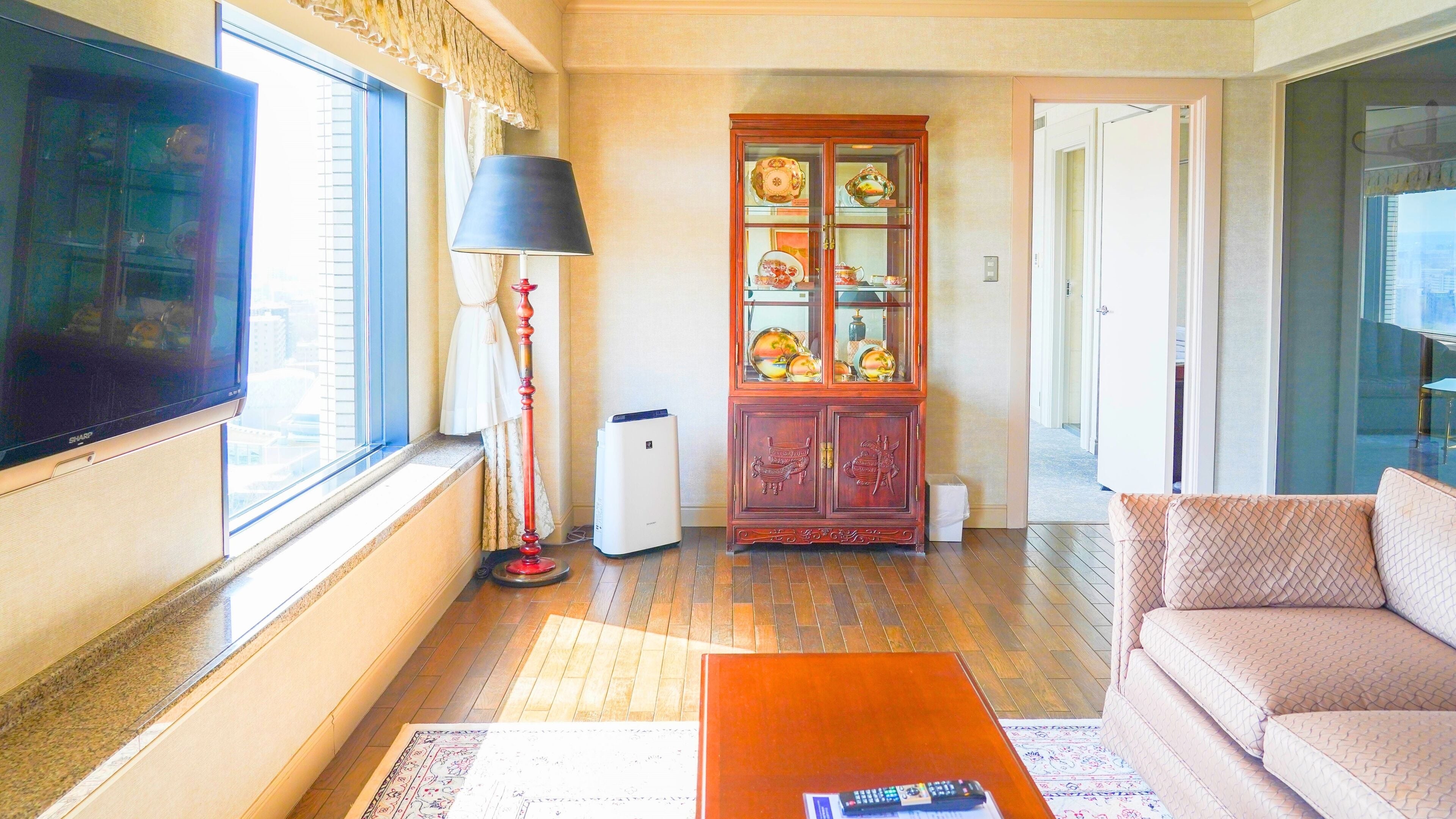Una sala de estar con un televisor de pantalla plana, una lámpara de pie roja, un mueble de madera con puertas de cristal, un purificador de aire blanco y un sofá color beige.