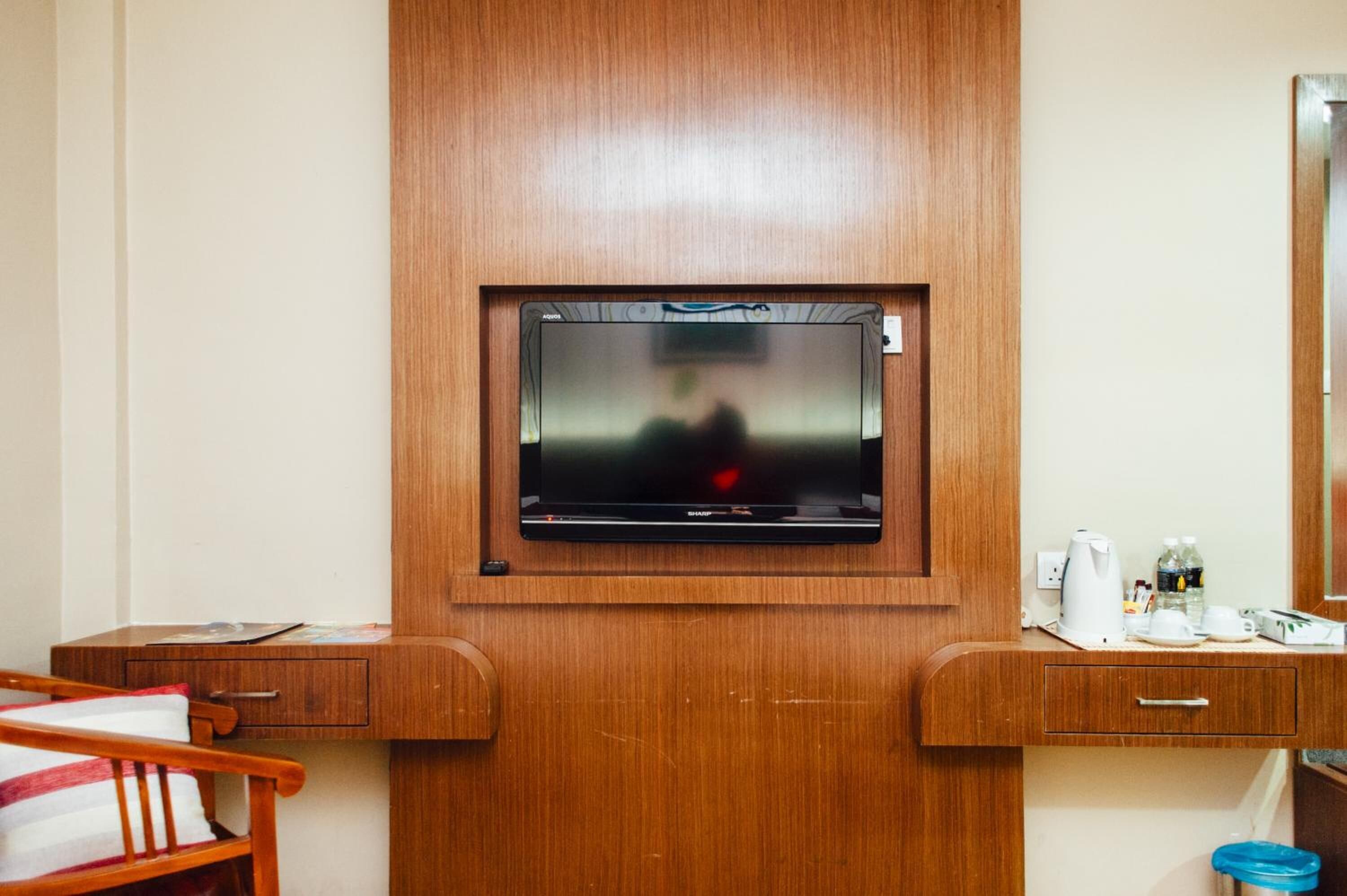 Habitación de hotel con un mueble de madera, un televisor montado en la pared, una mesita con hervidor, tazas y una botella de agua.