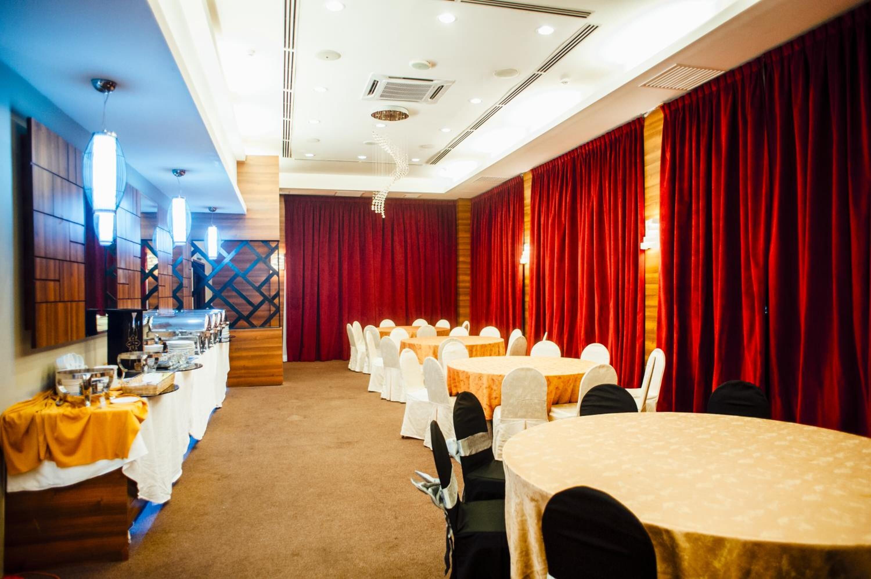 Un salón de conferencias con mesas redondas y sillas blancas, cortinas rojas y un montaje tipo buffet en un lateral.