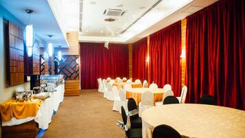 Un salón de conferencias con mesas redondas y sillas blancas, cortinas rojas y un montaje tipo buffet en un lateral.